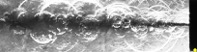 単一オリフィスによるキャビテーション噴流で捉えられた流れ場の合成画像（右端のノズル出口中央から左へ伸びる影が気泡群、明るい円弧状の波紋が気泡運動による生成衝撃波である。画像幅(約8 cm)にわたり、衝撃波の発生が確認できる。）